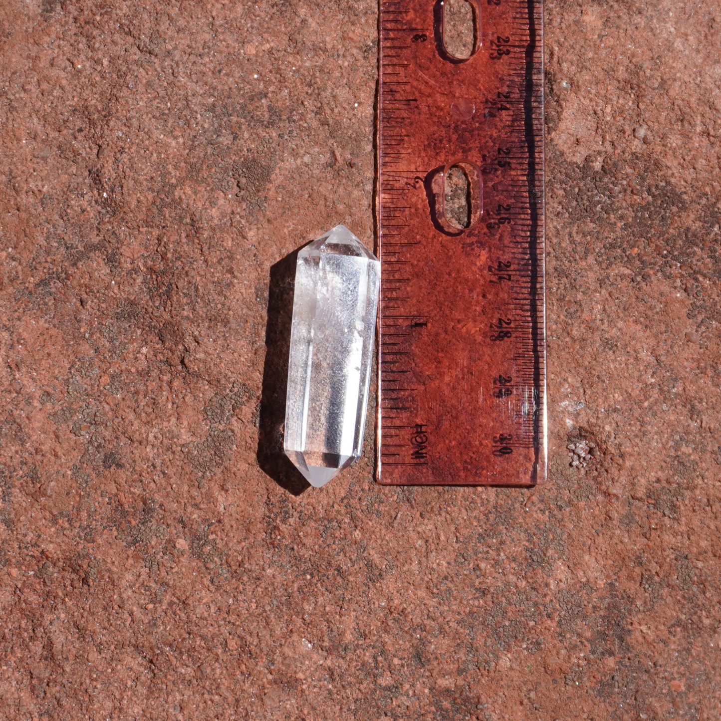 Measured clear quartz crystal beside ruler on sandstone, approximately 1.5 inches for accurate sizing