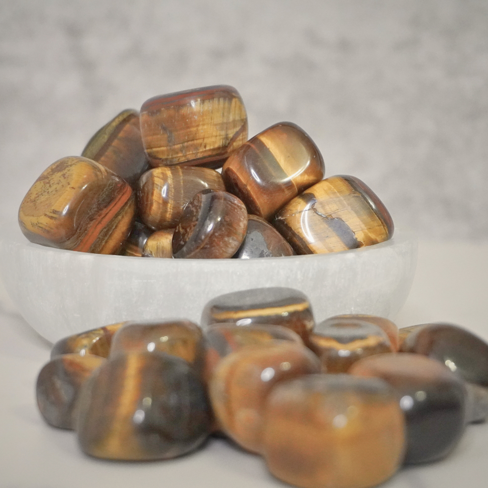 Close-up of polished Tiger's Eye tumbles in a white bowl, emphasizing the stone's grounding energy and reflective surface.