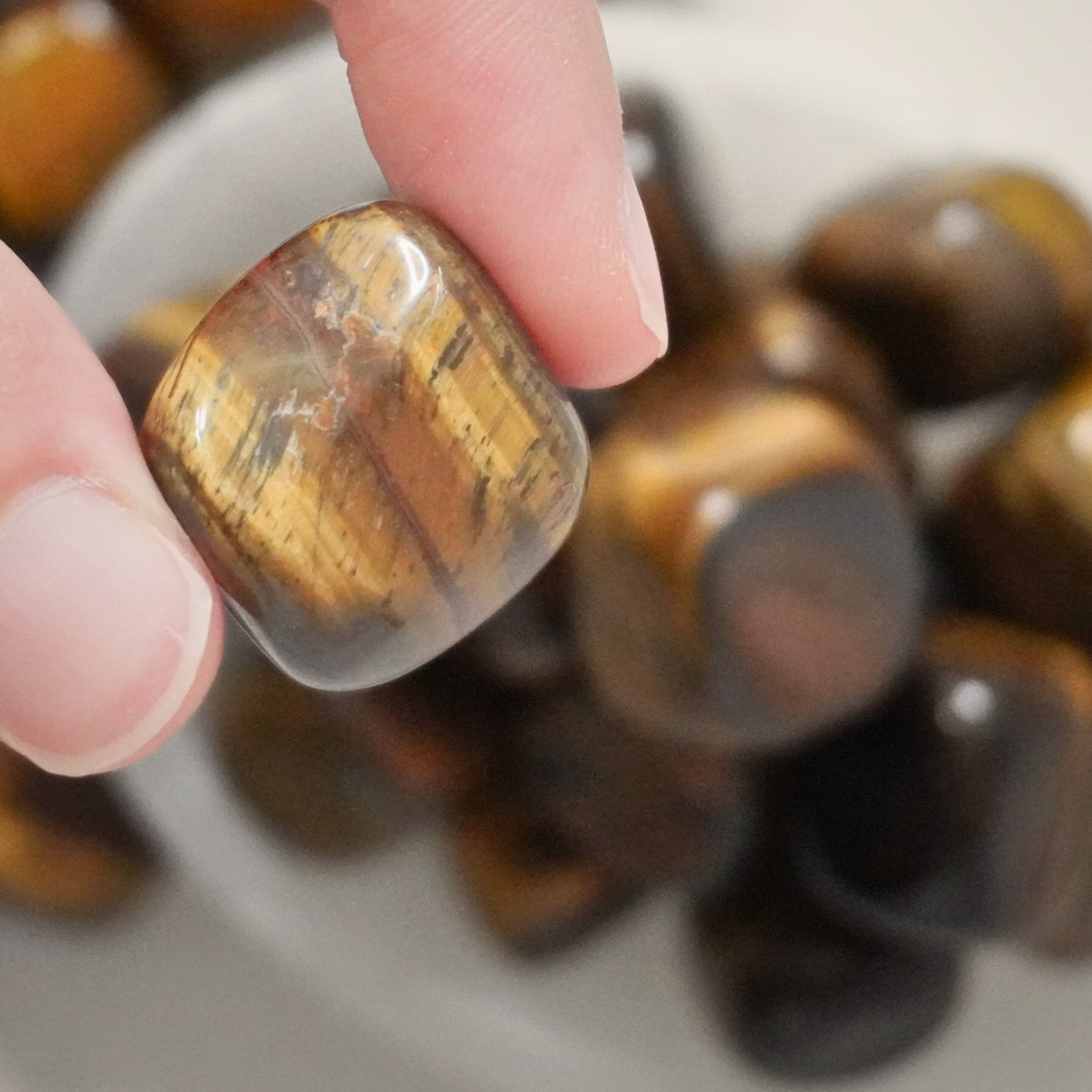 A hand selecting a polished Tiger's Eye tumble from a scattered group, showcasing the stone's grounding and balancing properties.