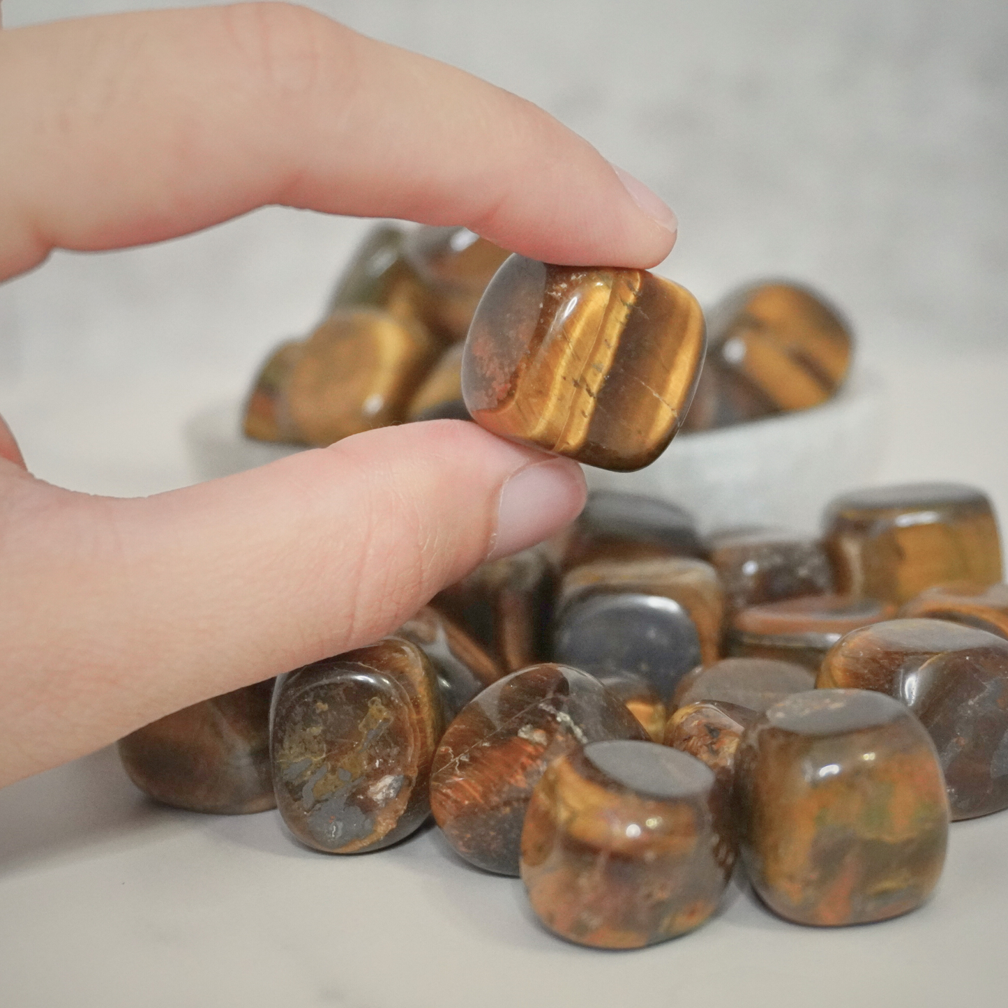 Tiger's Eye tumbles arranged in a bowl, with a hand gently touching them, emphasizing the stone's protective and stabilizing energy.